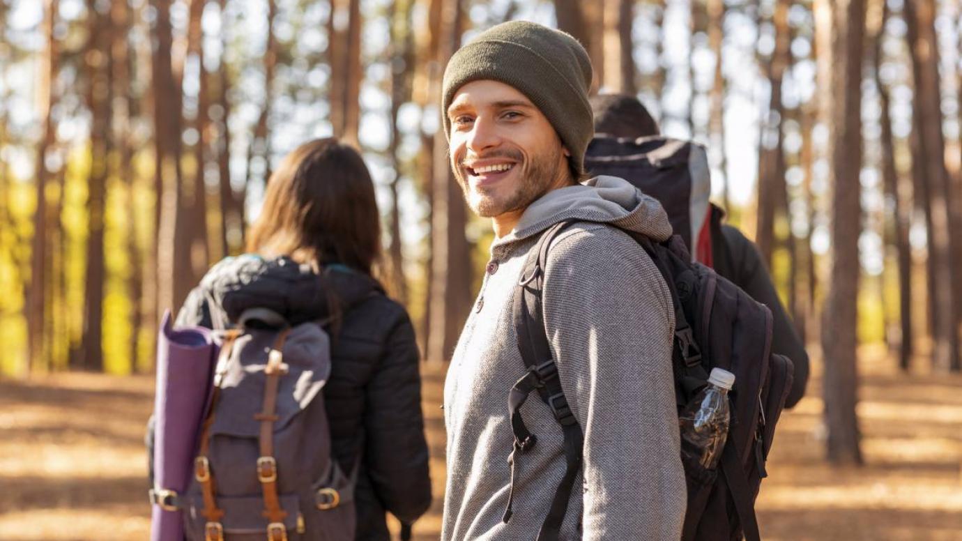 Smiling young man backpacker hiking with friends at forest, looking at camera, back view