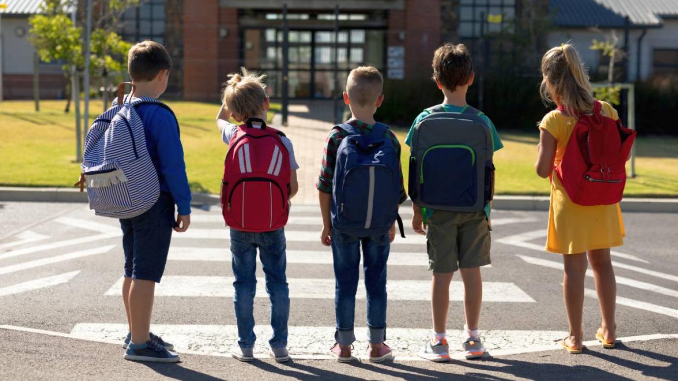 Rear view of a group of five schoolchildren carrying rucksacks waiting at a pedestrian crossing on a sunny day