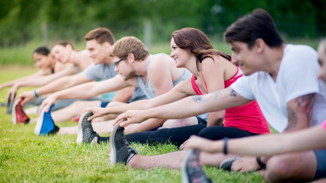 A multi-ethnic group of young adults are stretching together after a run. They are wearing athletic clothing and tennis shoes and are sitting in a row on the grass at the park.