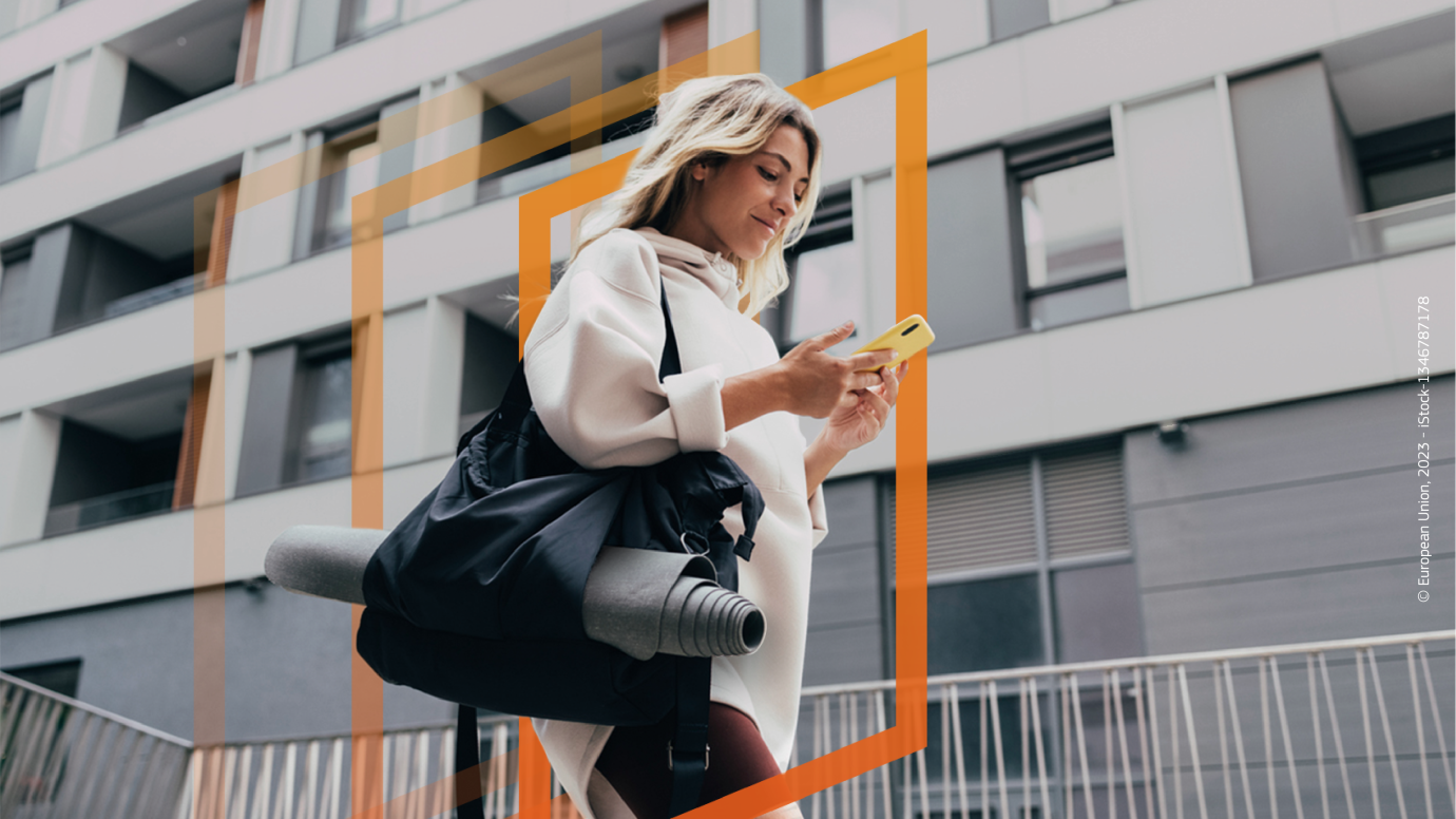 A girl walking on the street looking at her phone, carrying a gym bag and a yoga mat