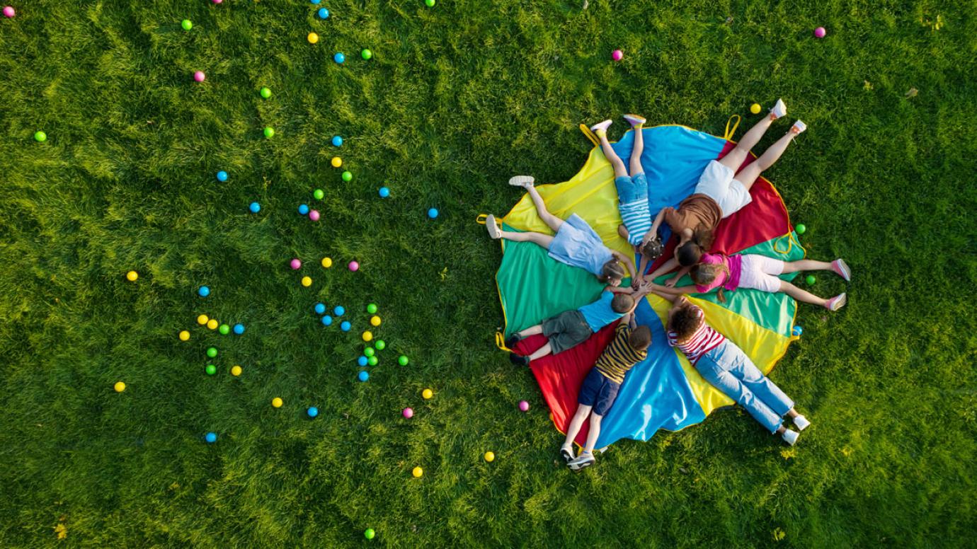 A group of children with their teacher holding hands on a rainbow playground parachute in a park, top view.